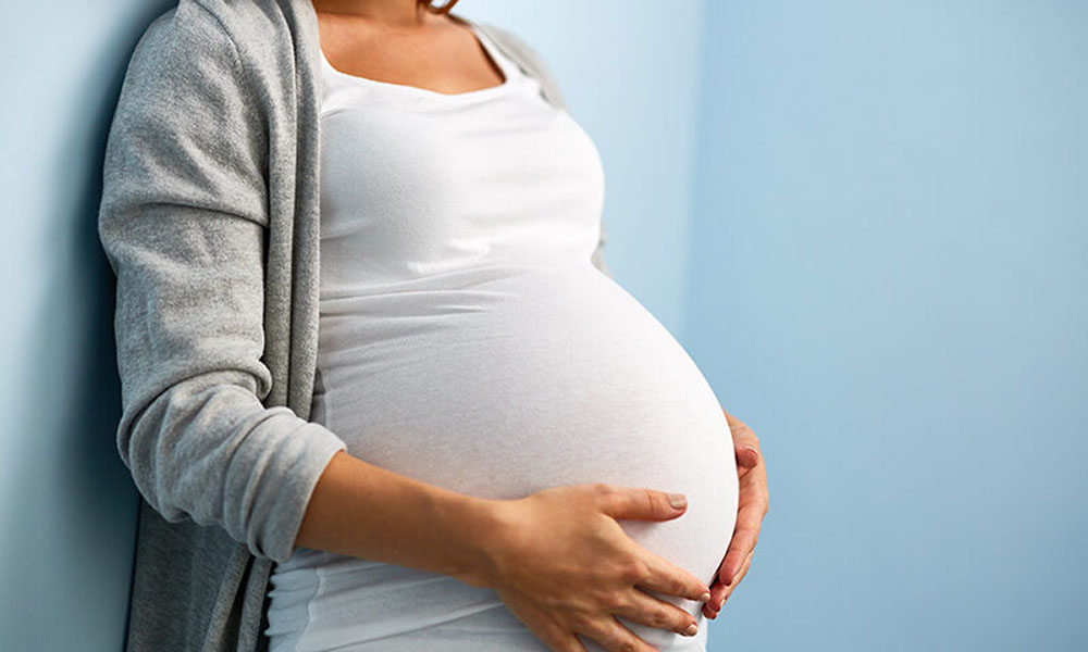 Mid-section portrait of unrecognizable woman during last months of pregnancy holding her big belly gently standing against wall in blue room